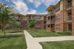 A view of an apartment community with several multi-story buildings, featuring balconies and landscaped greenery. The pathway in the foreground leads through a neatly maintained lawn, surrounded by trees and shrubs under a partly cloudy sky.