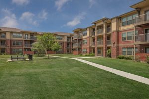A view of a residential apartment community featuring multiple buildings with balconies. The well-maintained green lawn has a pathway running through it, and there's a bench and a trash bin visible in the foreground. The sky is partly cloudy, creating a pleasant atmosphere.