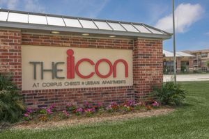 Sign for "The Icon at Corpus Christi Urban Apartments" with red lettering on a beige background. Surrounded by well-maintained landscaping featuring colorful flowers and green grass, under a clear blue sky. The building in the background is a multi-level apartment community.