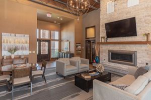 A modern living room with a cozy atmosphere featuring a stone fireplace, comfortable seating, and a round dining table. Large windows allow natural light, while decorative artwork and plants add a touch of style. A flat-screen TV is mounted above the fireplace, and warm lighting enhances the inviting space.