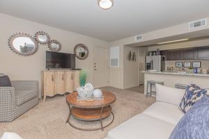 A cozy living room featuring a beige carpet, a light-colored sofa adorned with decorative pillows, and a round wooden coffee table. There's a wooden side table with decorative items, a television on a stand, and wall mirrors with gear designs. The room opens to a kitchen area with dark cabinetry.