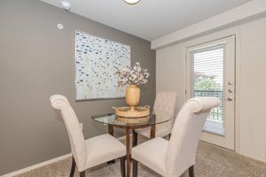 Cozy dining area featuring a round glass table with a wicker basket and a decorative vase filled with branches. Two upholstered chairs with a neutral color scheme are placed around the table, against a backdrop of a gray wall and a large artwork depicting abstract blue shapes. A door with blinds leads to an outdoor space.