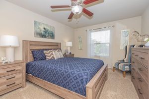 A cozy bedroom featuring a wooden bed frame with a blue patterned comforter, two nightstands, and a dresser. The room has tasteful artwork on the walls, a ceiling fan, and a large window with white curtains. A striped chair adds to the decor, creating a warm and inviting atmosphere.