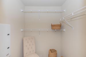 A well-organized closet featuring two white wire shelves against light-colored walls. A beige upholstered chair is positioned in the corner, alongside two woven baskets on the floor, adding a touch of warmth to the space. The closet appears tidy and functional, suitable for storage.