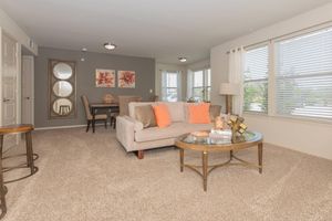 A cozy living room featuring a light beige sofa with orange pillows, a glass coffee table, and a decorative centerpiece. The walls are painted gray, and there are two windows with sheer curtains allowing natural light. A round mirror and artwork with floral designs adorn the walls, along with a small dining area in the background.