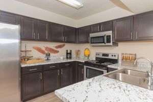 A modern kitchen with dark wooden cabinets, granite countertops, stainless steel appliances, and a double sink. Decor includes a basket with kitchen utensils and a display of oranges on the countertop. The overall design is sleek and contemporary, with neutral colors and good lighting.