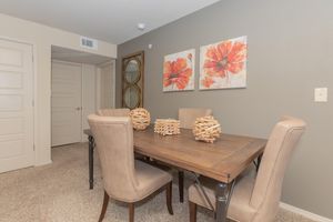 A modern dining area featuring a wooden table surrounded by four beige upholstered chairs. On the table, there are decorative wooden pieces arranged in two clusters. The walls are adorned with two vibrant floral paintings, and a mirror is visible in the background against a neutral-colored wall. A door leads to another room.