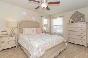 A cozy bedroom featuring a light-colored wooden bed with a patterned quilt, two bedside lamps, a beige dresser, and a large window with sheer curtains. The wall displays decorative flowers in pink and white. The room has a soft, inviting ambiance with neutral tones and carpet flooring.