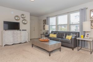 A cozy living room featuring a dark leather sofa with yellow accent pillows, a wooden coffee table, and a decorative bowl. Opposite is a light-colored TV stand with a television, flanked by two lamps. The room has beige carpet, gray curtains, and wall mirrors that enhance the space's brightness.
