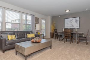 A cozy living room with a dark leather sofa adorned with yellow accent pillows, a wooden coffee table, and a decorative bowl. On the wall is a modern art piece featuring a dog. Natural light filters through large windows with blinds, while a dining area with a wooden table and chairs is visible in the background.