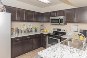 Modern kitchen featuring dark wood cabinets, stainless steel appliances including a microwave and oven, and a granite countertop. The space is adorned with decorative items like yellow dishes and framed floral art. There are two sinks and a stylish backsplash, creating a contemporary and inviting atmosphere.