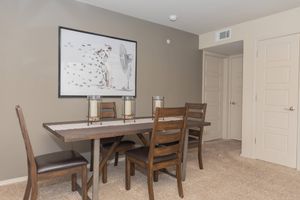 Dining area featuring a wooden table with four chairs, a neutral-toned wall, and a large framed artwork of a dog by a fan. The setting includes a light-colored table runner and two decorative lanterns. A door and a closet add functionality to the space. Soft carpet underfoot completes the cozy atmosphere.