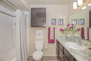 A modern bathroom featuring a white bathtub with a patterned shower curtain, a stylish sink with a granite countertop, dark wooden cabinets, and decorative framed artwork. Fresh flowers adorn the counter, and a motivational sign reads "Keep Life Simple." The space is well-lit and organized.