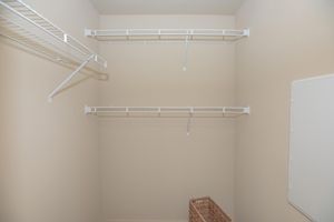 Empty closet with beige walls and white wire shelving. Two shelves are mounted on the walls, while one lower shelf is partially visible. A woven basket is placed on the floor, adding a touch of texture. The overall space is clean and minimalistic, suited for storage needs.