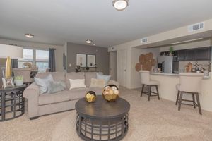 A cozy living room featuring a beige sofa adorned with decorative pillows, a round coffee table with geometric decor, and a dining area with barstools. The walls are painted in neutral tones, and there are decorative wall hangings. Natural light enters through the windows, enhancing the inviting atmosphere.