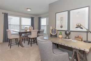 A modern, well-lit living space featuring a dining area with a table and chairs, and a desk with a stylish lamp. The walls are adorned with framed artwork, and there are decorative items on the table. Soft gray carpet and window treatments add warmth to the room.
