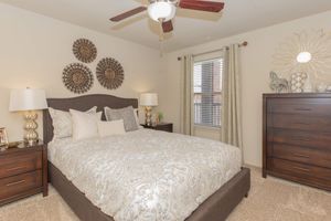 A cozy bedroom featuring a neatly made bed with decorative pillows, two stylish bedside tables with lamps, and a brown dresser. Three circular wall decorations hang above the bed, and a ceiling fan is visible. Natural light enters through a window with blinds. The carpeted floor adds warmth to the space.