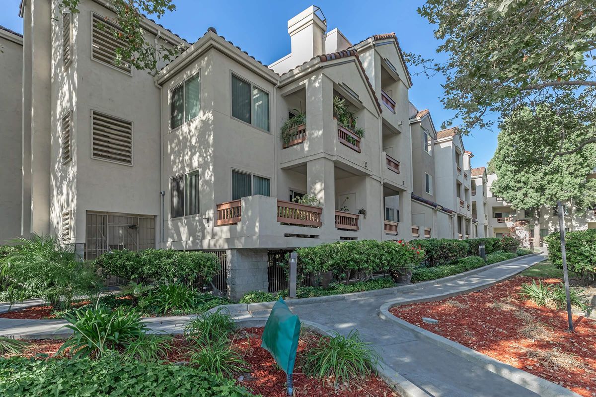 A well-maintained multi-story apartment building featuring a landscaped pathway, greenery, and balconies with planters. The architecture includes beige walls and a tile roof, with a clear blue sky in the background. Paths are lined with red mulch and decorative plants, creating an inviting residential environment.