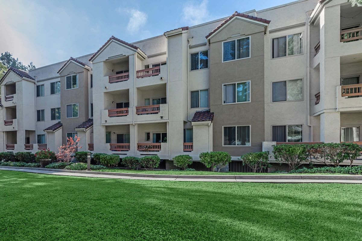 A modern apartment building with multiple floors, featuring balconies and large windows. The exterior is painted in light beige with dark accents. In front, there is a well-maintained green lawn and landscaped shrubs, creating a pleasant outdoor space. The scene is bright and inviting.