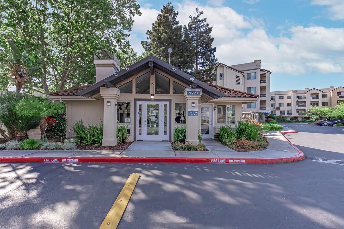 Entrance of a modern residential building featuring a peaked roof, large glass doors, and surrounding greenery. A parking lot is visible in front with clear markings, and a backdrop of tall trees and multi-story apartments. The scene appears bright and welcoming.