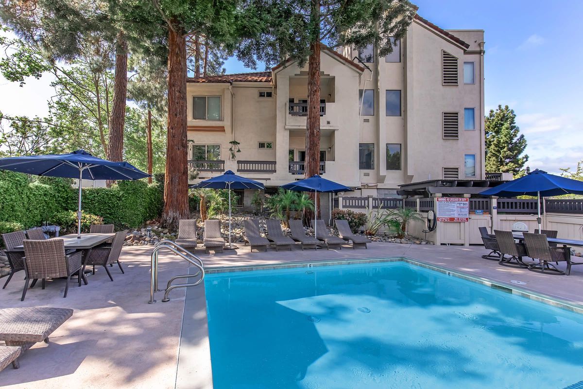 A serene outdoor pool area with a clear blue pool surrounded by lounge chairs and umbrellas. In the background, a multi-story building with balconies and tall trees adds to the tranquil atmosphere. Perfect for relaxation and leisure on a sunny day.