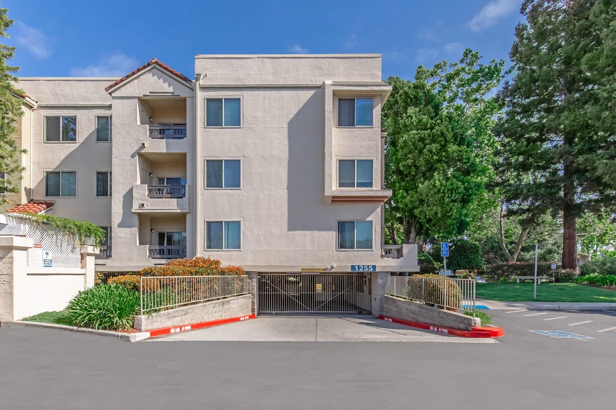 Three-story apartment building with a light-colored exterior, featuring balconies on the upper floors. There's a gated entrance with a marked parking area in front. Lush greenery surrounds the property, with trees and shrubs enhancing the landscape. The sky is bright and clear.