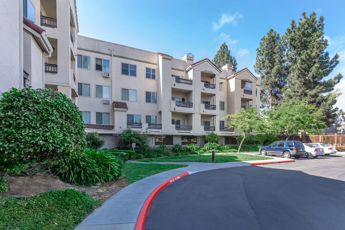 A multi-story residential building with a beige exterior, featuring balconies and landscaped greenery, including shrubs and trees. A paved driveway curves around the property, with a parked car visible. The scene is set under a clear blue sky.
