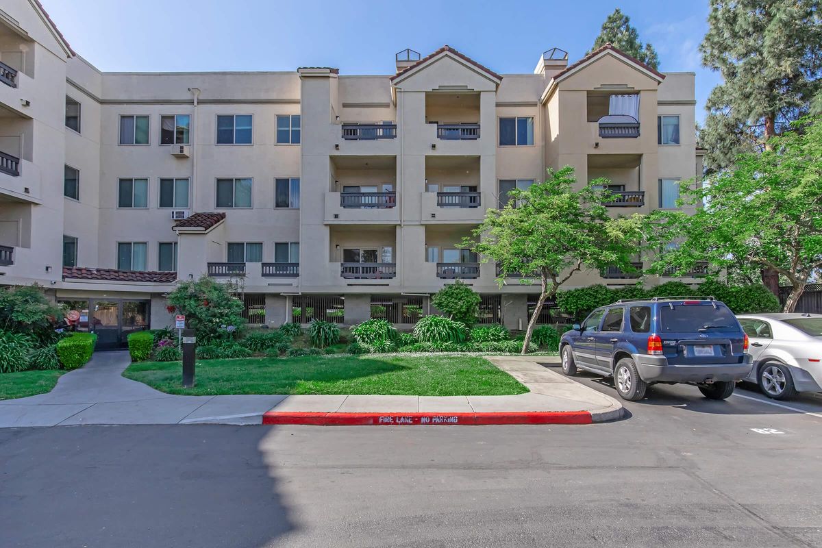 A multi-story residential building with a light beige exterior, featuring balconies on the upper floors. The landscaped area includes trees and shrubs. A parking lot is visible in the foreground with a silver SUV parked, and a red curb marked "FIRE LANE – NO PARKING." Clear blue sky in the background.