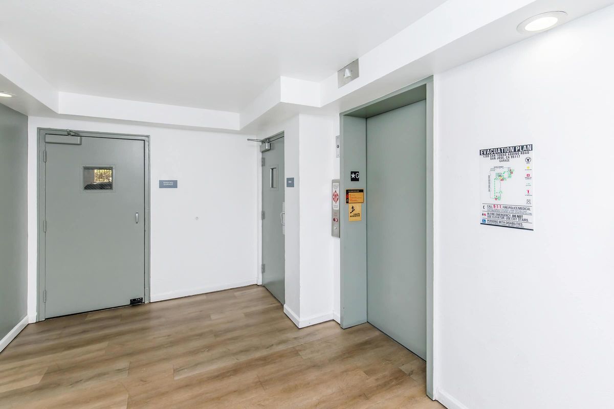 Interior view of a modern building hallway with light-colored walls and wooden flooring. Features include an elevator door, a door labeled for use, and a wall-mounted directory map. Soft lighting enhances the spacious appearance of the corridor.