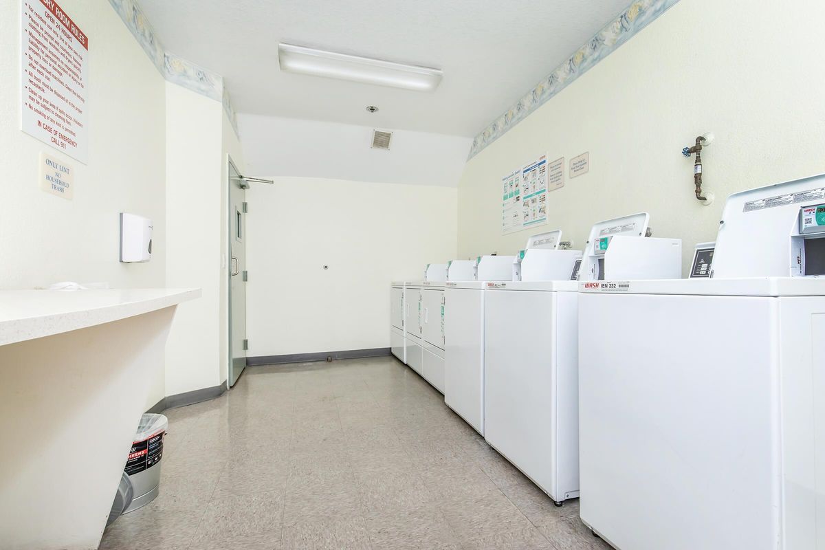 A clean and well-lit laundry room featuring several white washing machines aligned against the wall. There is a counter space on the left side and a door at the far end. Fresh, neutral-colored walls and a tiled floor contribute to the bright and tidy atmosphere.