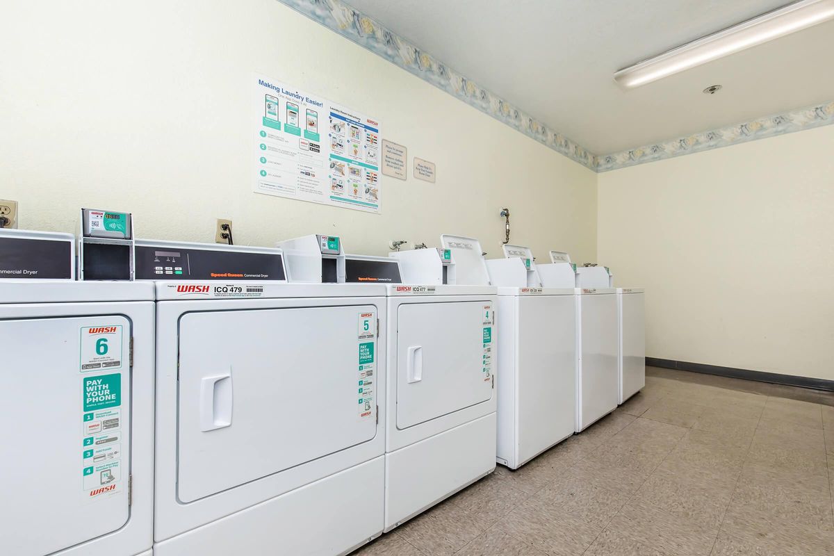 A laundry room containing several white washing machines and dryers lined up against a wall. The room has a light-colored, clean appearance with a bulletin board and signs on the wall providing laundry instructions. The floor is tiled, and there is overhead lighting.
