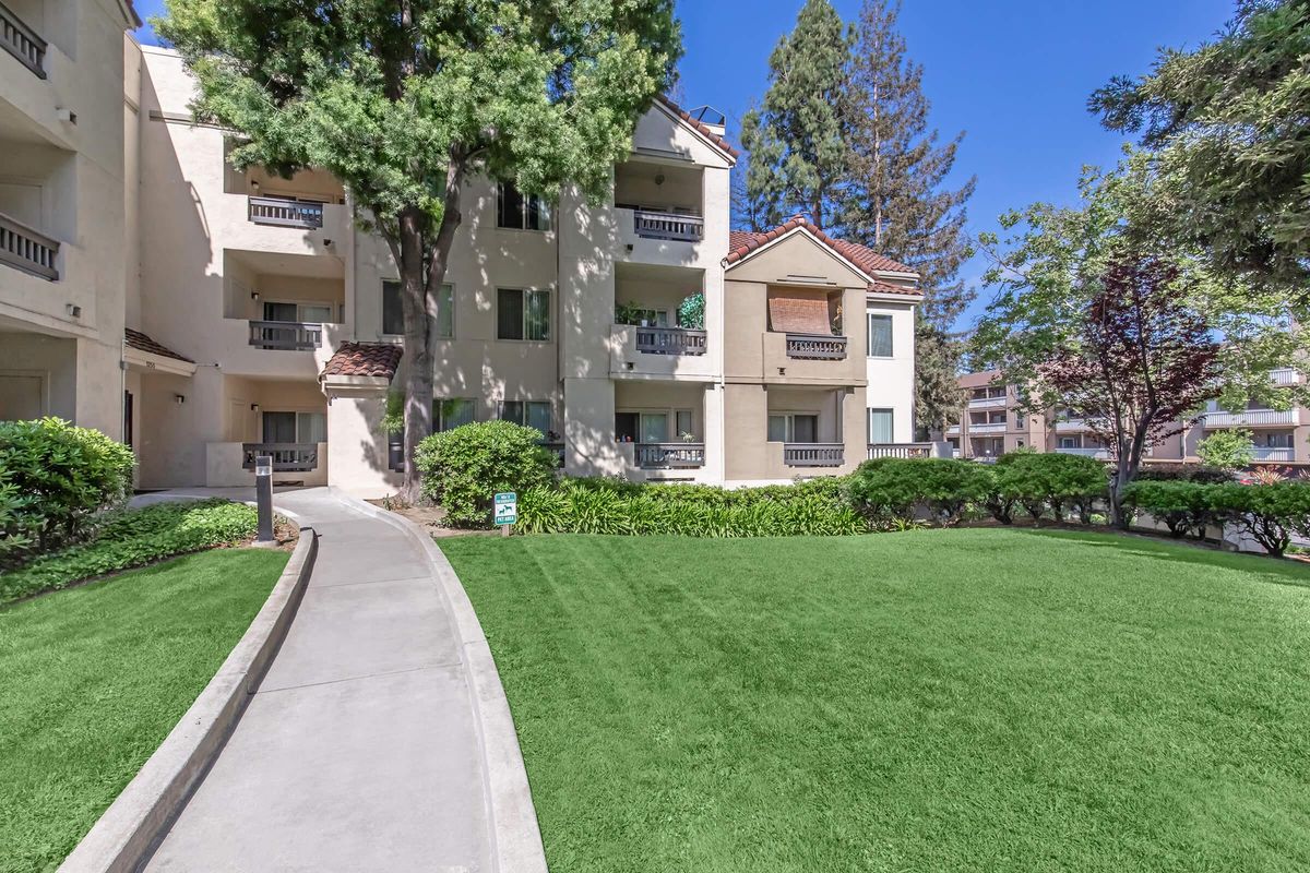 A well-maintained walkway leads through a lush green lawn towards an apartment building surrounded by trees and shrubs. The building features multiple balconies and large windows, with a clear blue sky overhead.
