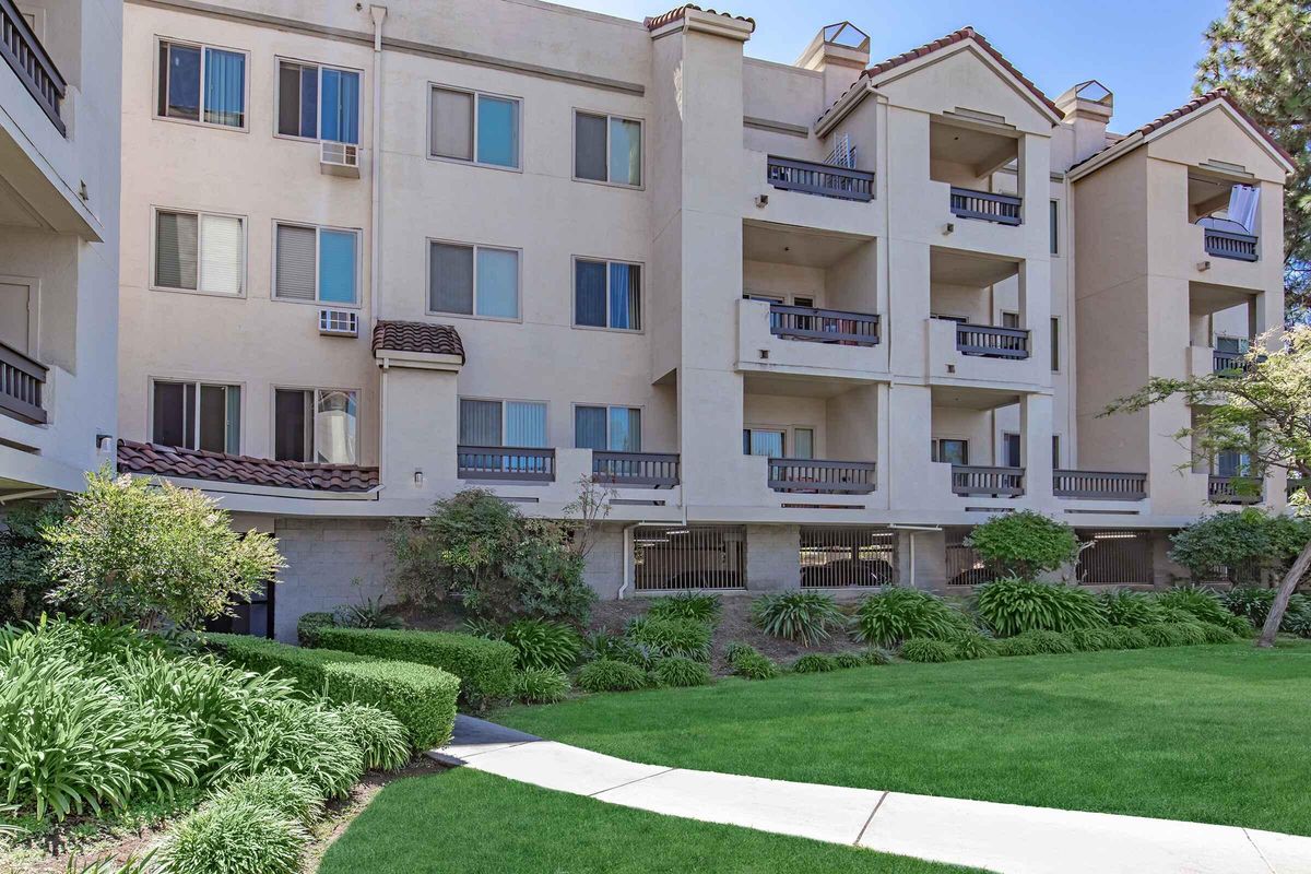 A residential building with multiple floors featuring balconies. The exterior is light-colored with a tiled roof. There is a well-maintained green lawn in the foreground with lush plants and shrubs. Windows are visible on each floor, and some balconies have potted plants. Sunlight brightens the scene.