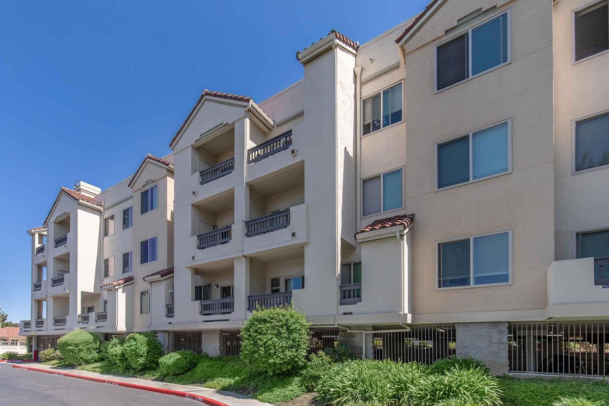 A row of modern apartment buildings with beige exteriors and terracotta roofs. The structure features multiple balconies, large windows, and surrounding green shrubs. The sky is clear and blue, indicating a sunny day.