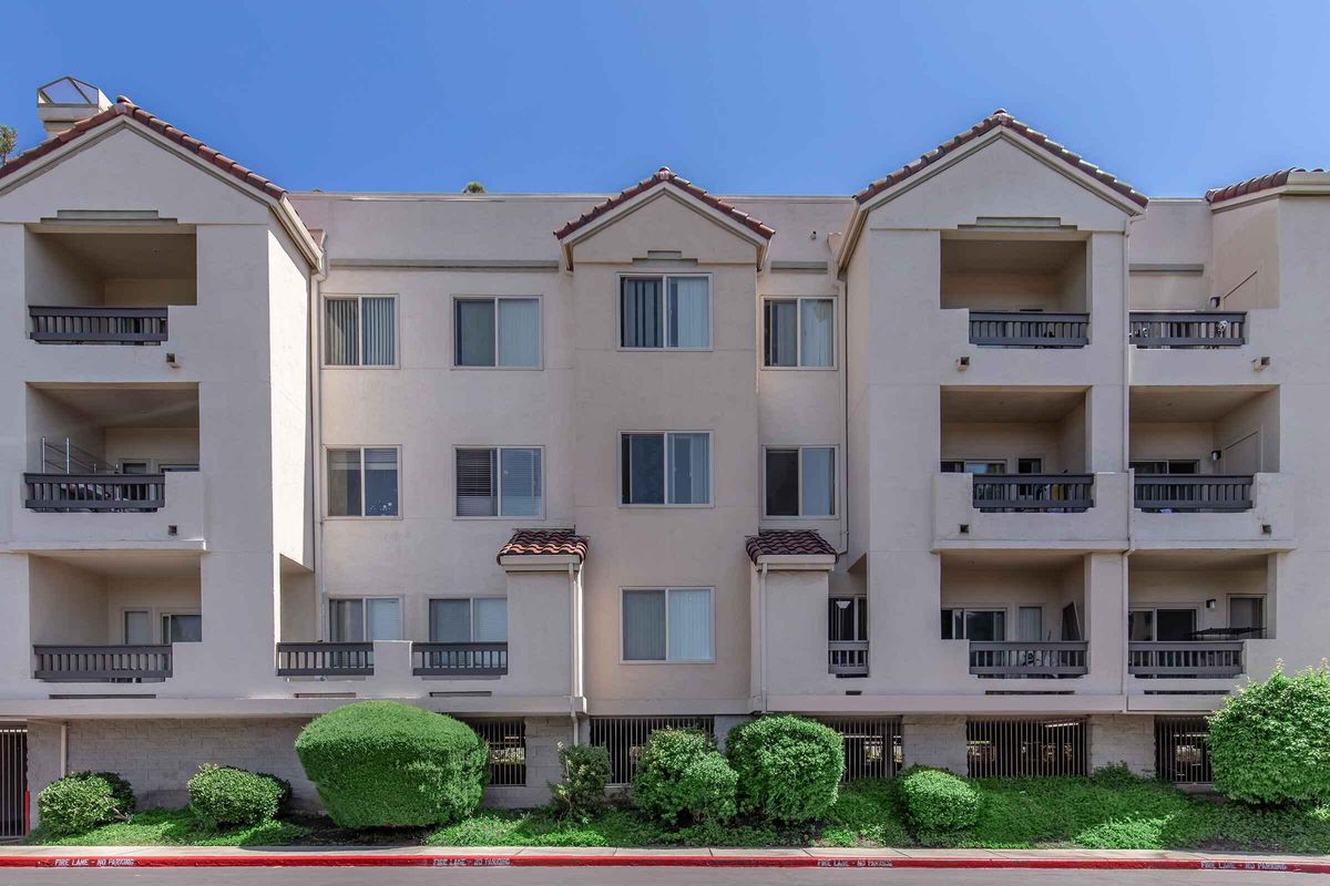 A three-story apartment building with a beige exterior, featuring balconies and large windows. The building is flanked by neatly trimmed green bushes. The sky is clear and blue, creating a bright background.