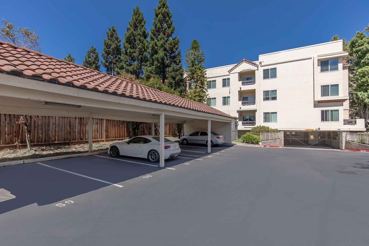 A parking lot with covered spaces and two parked cars, set against a backdrop of a four-story residential building with a red-tiled roof and surrounding trees. The lot is well-maintained, and the sky is clear, indicating a sunny day.