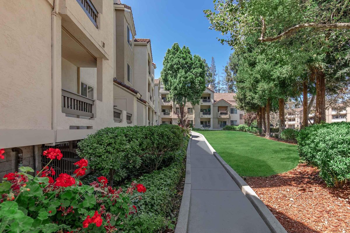 A landscaped path lined with vibrant red flowers leads through a residential area. The path is flanked by greenery and trees, with beige apartment buildings visible in the background under a clear blue sky. The scene conveys a peaceful, inviting atmosphere.