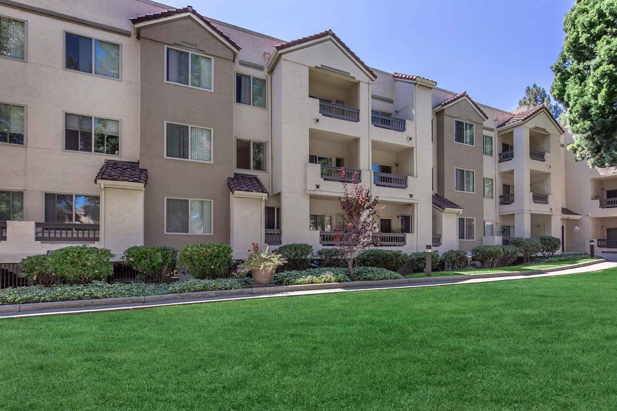 A well-maintained apartment community featuring multiple beige buildings with balconies, surrounded by manicured lawns and shrubs. The scene is set on a bright, sunny day, highlighting the greenery and inviting atmosphere of the outdoor space.