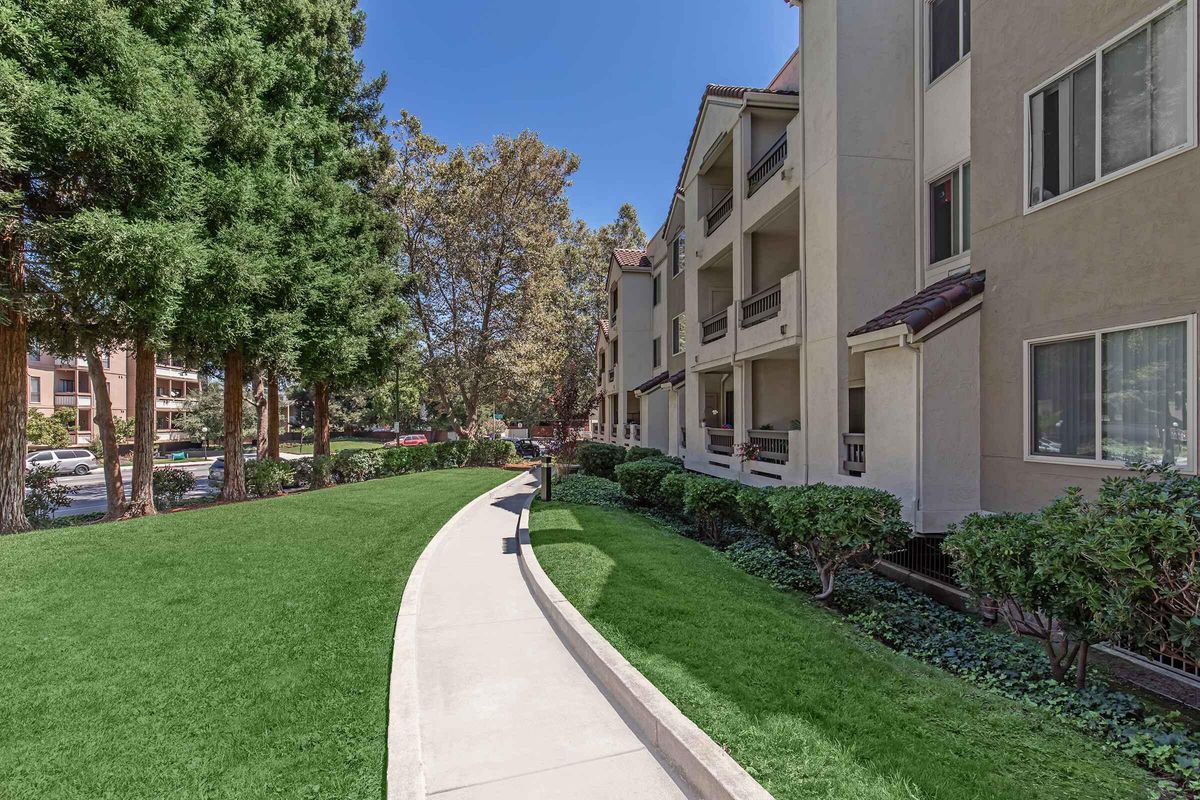 A landscaped pathway curves through a grassy area, flanked by neatly trimmed bushes and trees. In the background, residential apartment buildings are visible, with balconies and windows. The scene is set under a clear blue sky, giving a warm and inviting atmosphere.