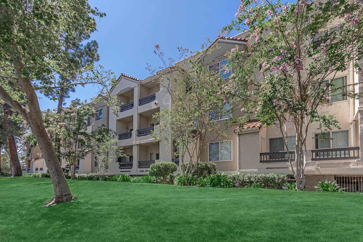 A modern apartment building surrounded by lush green grass and trees, with blooming flowers. The building features balconies and a well-maintained exterior under a clear blue sky.