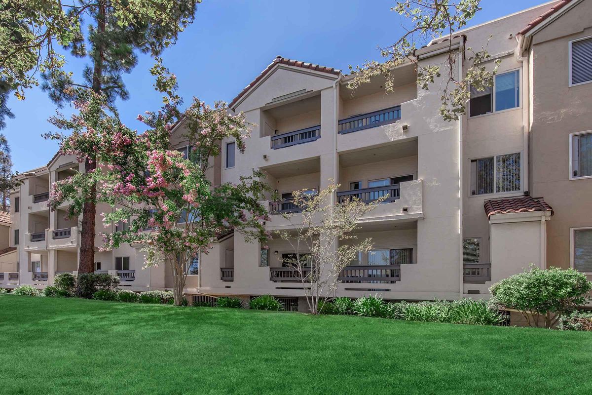A three-story apartment building with a Mediterranean architectural style, featuring balconies and large windows. The building is surrounded by lush green grass and flowering trees under a clear blue sky, creating a serene and inviting atmosphere.