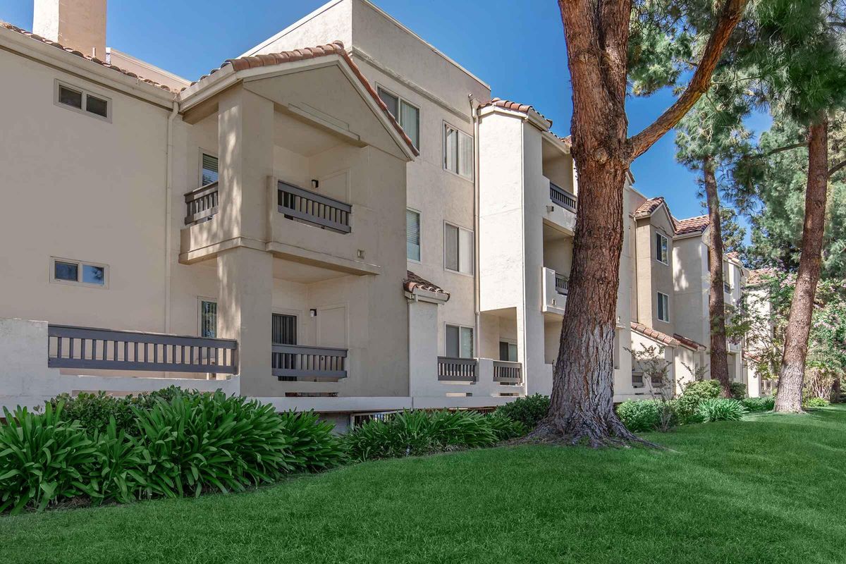 A view of a residential apartment community featuring multiple white stucco buildings with balconies. Lush green grass and shrubs line the foreground, and tall trees provide shade nearby. The architecture reflects a modern design with Spanish influences, set against a clear blue sky.