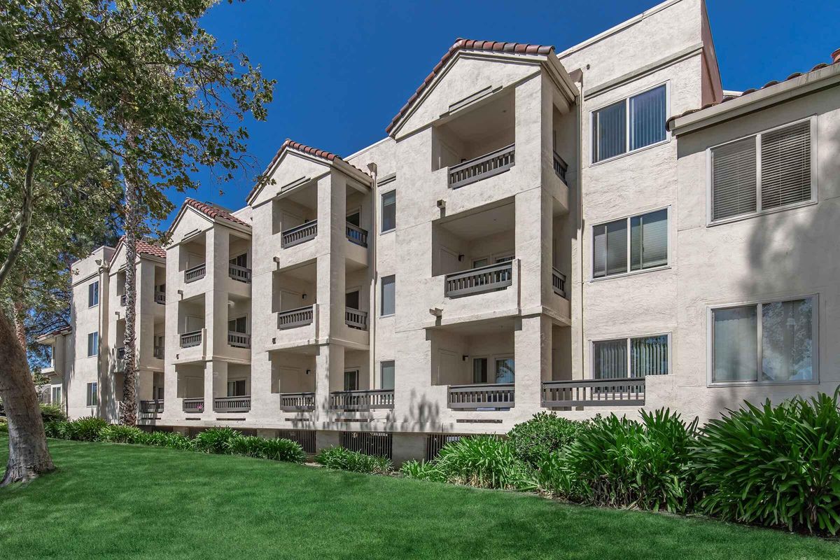 A two-story apartment building with a stucco exterior and multiple balconies. The building is set against a bright blue sky and features green grass and shrubs in the foreground. The architecture includes arched windows and a tiled roof, conveying a modern yet classic style.