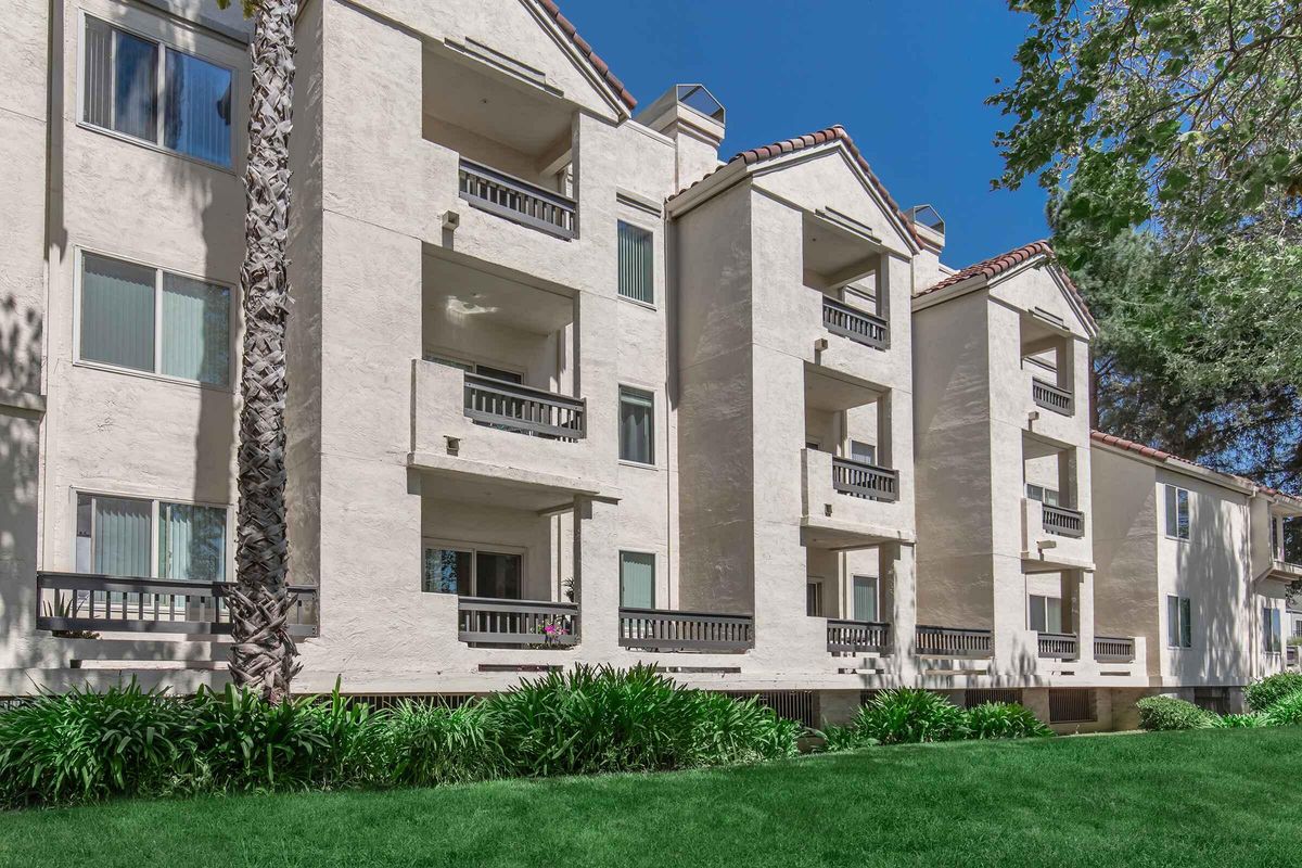 Three-story residential building with light-colored stucco walls, featuring multiple balconies and large windows. The surrounding area has lush green grass and landscaping, including palm trees. The clear blue sky creates a bright, inviting atmosphere.