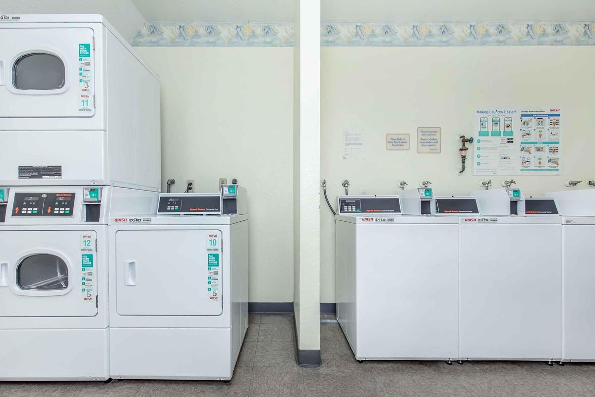 A clean, well-lit laundry room featuring stacked washing machines and dryers on the left, and row of front-loading washers with coin slots on the right. The walls are painted a light color with a decorative border, and there are informative signs and instructions displayed nearby.