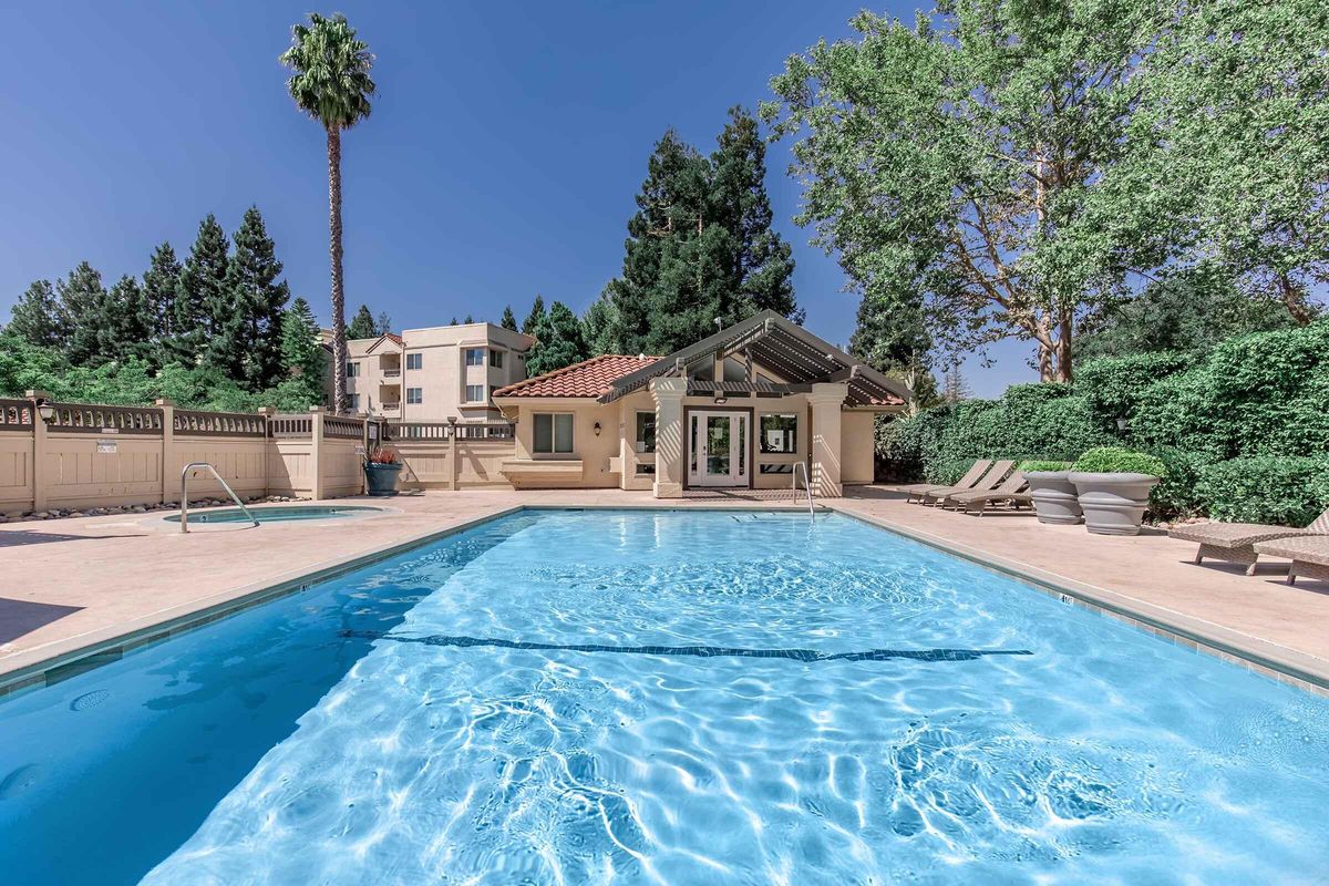 A clear blue swimming pool surrounded by palm trees and lush greenery. In the background, a small building with a tiled roof and large windows overlooks the pool area, which features lounge chairs and a serene atmosphere under a bright blue sky.