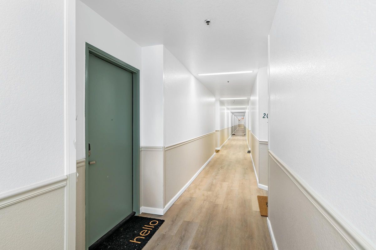 A long, well-lit hallway with white walls and a light wooden floor. Two doors are visible, one green and one white, with a welcome mat saying "hello" in front of the green door. The hallway has a clean, modern aesthetic with minimal decor.