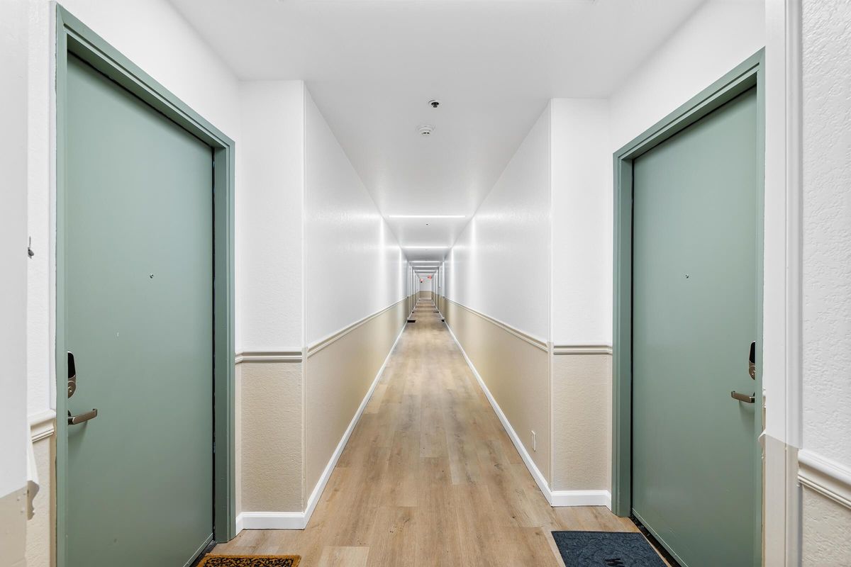 A long, well-lit hallway with two green doors on either side. The walls are painted white, and there is a wooden floor. Decorative mats are placed in front of the doors, leading down the straight corridor toward a distant perspective.