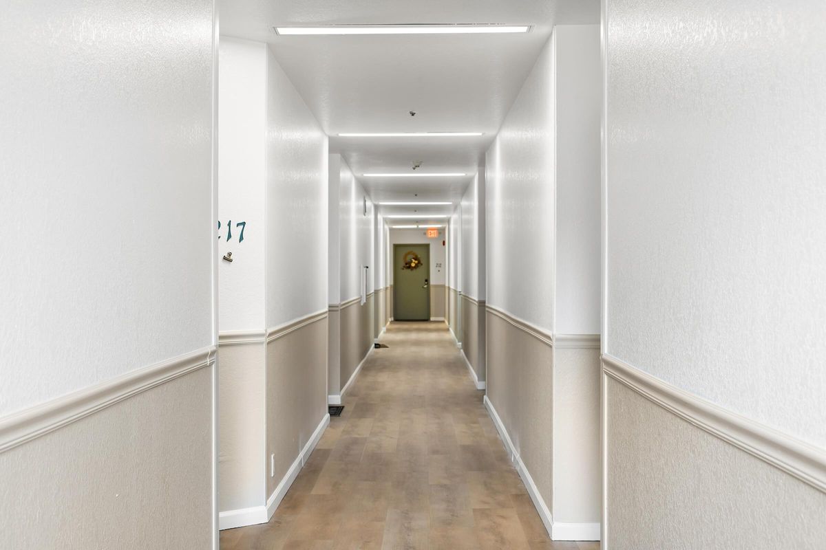 A well-lit corridor with white walls and light-colored flooring, featuring a decorative wall element at the end. Doors line either side, with a visible door marked "217." The lighting is uniform, creating a spacious and clean atmosphere.