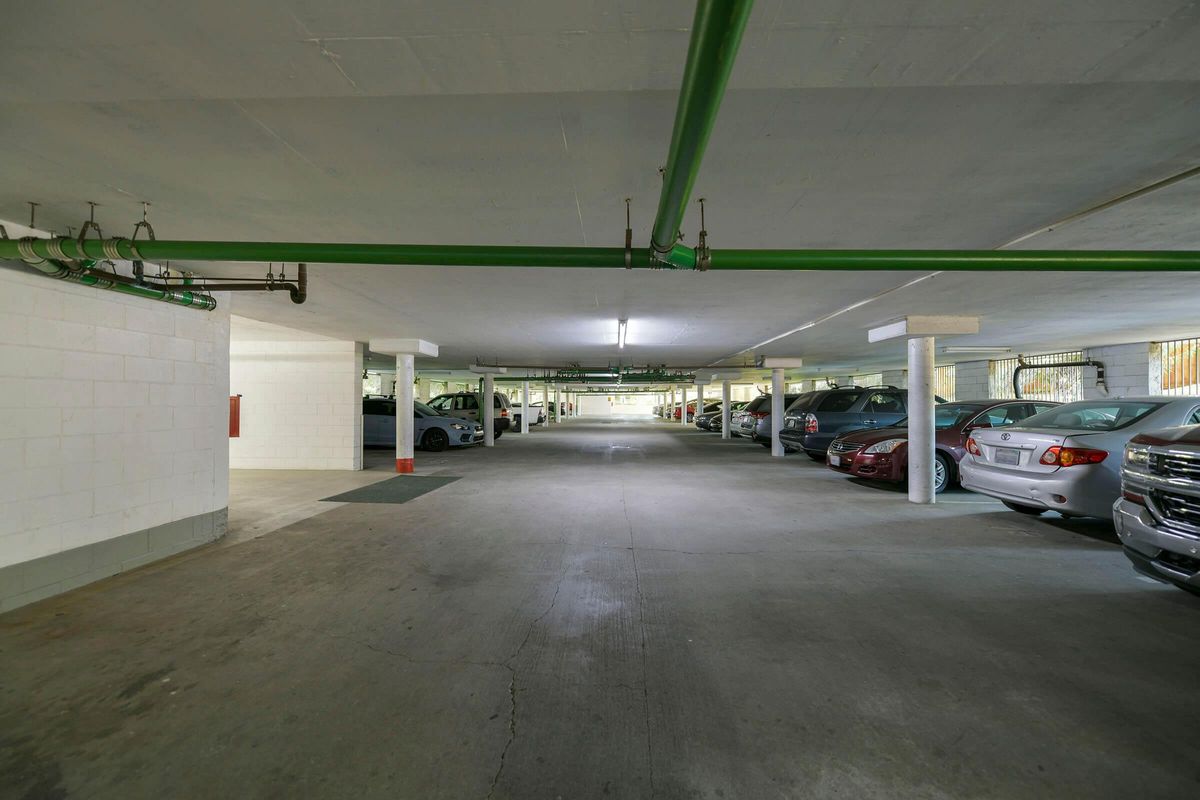 A spacious, well-lit underground parking garage featuring several parked cars on both sides. The concrete floor is visible, along with green pipes running along the ceiling and rows of white columns supporting the structure. The far end of the garage is dimly lit, leading to an exit.